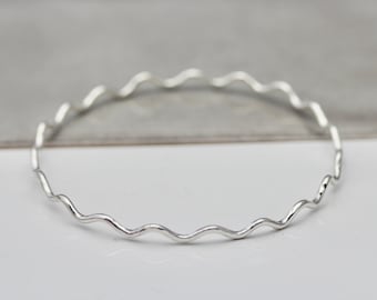 May include: A thin, circular bangle bracelet crafted from polished silver-toned metal. The band features a continuous, repeating wavy or rippled pattern throughout its entire circumference, creating a delicate and textured appearance.