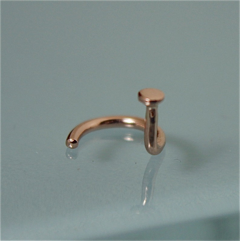 May include: A silver nose ring with a small, round top. The ring is made of metal and has a curved shape.