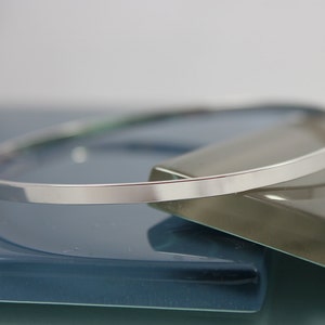 May include: A minimalist silver bangle bracelet. The bracelet has a flat, rectangular profile and a smooth, polished surface. The jewelry is displayed on a reflective surface with blue and gray tones.