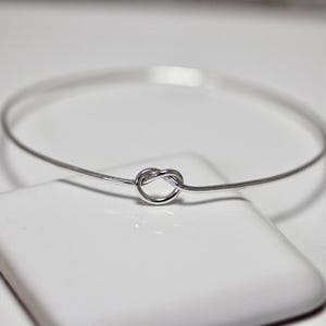 May include: A delicate silver-toned bangle bracelet with a knot design at the center. The bracelet is thin and circular, with a simple, elegant aesthetic. The knot detail adds a unique touch to the minimalist jewelry piece.