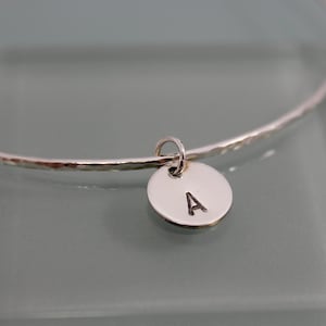 May include: A silver bangle bracelet with a hammered finish and a small silver charm with the letter "A" engraved on it.