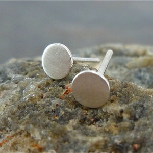 May include: A pair of silver stud earrings with a round, flat design. The earrings are simple and minimalist.