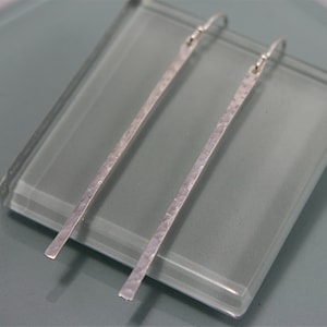 May include: A pair of long, silver earrings with a hammered texture. The earrings are hanging from a glass surface.