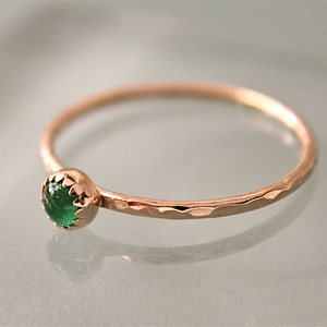 May include: A delicate gold ring with a hammered band and a bezel-set, round, emerald-green gemstone. The ring's setting has a scalloped edge, and the band has a textured, hammered finish. The ring is a piece of jewelry.