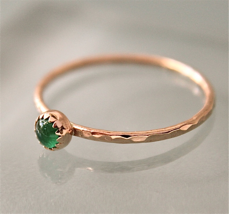 May include: A delicate gold ring with a hammered band and a bezel-set, round, emerald-green gemstone. The ring's setting has a scalloped edge, and the band has a textured, hammered finish. The ring is a piece of jewelry.