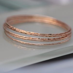 May include: A stack of four rose gold bangles with a hammered texture. The bracelets are thin and circular, reflecting light. The jewelry is displayed on a light-colored surface.