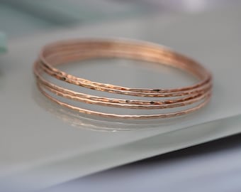 May include: A stack of four rose gold bangles with a hammered texture. The bracelets are thin and circular, reflecting light. The jewelry is displayed on a light-colored surface.