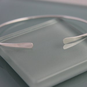 May include: A simple silver bangle bracelet with a flat, slightly curved design. The bracelet is open ended and has a smooth finish.