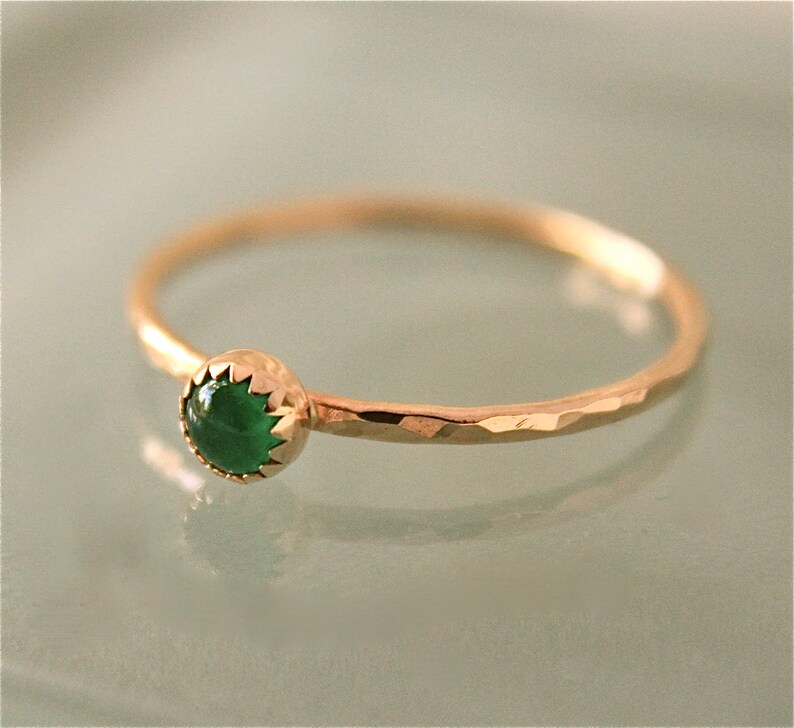 May include: A delicate gold ring with a hammered band and a bezel-set, round, emerald-green gemstone. The ring's band has a textured, faceted appearance, and the gemstone is the focal point.