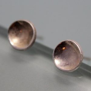 May include: A pair of silver stud earrings with a rounded, slightly concave surface. The earrings have a subtle, metallic sheen.