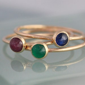 May include: Three gold rings with different coloured gemstones. The rings are stacked on top of each other. The top ring has a blue gemstone, the middle ring has a green gemstone, and the bottom ring has a red gemstone.