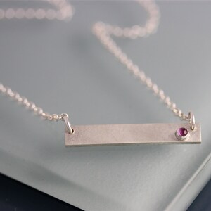Op de afbeelding: Een zilveren ketting met een rechthoekige hanger en een kleine roze edelsteen. De hanger is glad en heeft een geborstelde afwerking.