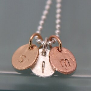May include: A silver chain necklace with three charms. The charms are round and made of different metals: gold, silver, and copper. The charms are engraved with the letters "S", "I", and "M".