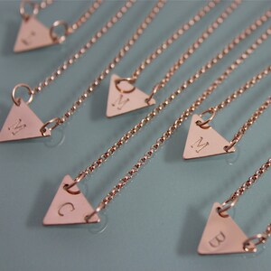 May include: Five gold triangle necklaces with different letters engraved on each. The letters are B, C, M, and two Ms.