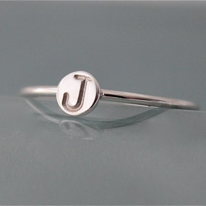 Op de afbeelding: Een zilveren ring met een kleine ronde bedel met de letter "J" erop gegraveerd.