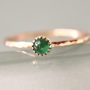 May include: A delicate gold ring featuring a vibrant green gemstone. The ring has a hammered texture on the band and a scalloped bezel setting around the round-cut emerald. The ring is a simple yet elegant piece of jewelry.