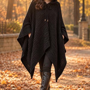 May include: A black hooded poncho featuring a textured knit pattern, a drawstring neckline with metallic aglets, and side pockets. The garment has an asymmetrical hemline and is worn over black trousers and black knee-high leather boots.