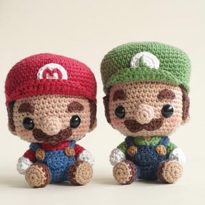 May include: Two small crocheted dolls representing characters with large black eyes, brown mustaches, and tan skin. One doll wears a red cap with a white M and red shirt, the other a green cap with a white L and green shirt. Both wear blue overalls.