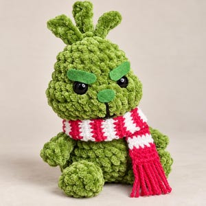 Op de afbeelding: Een groen pluche speelgoedfiguur gemaakt van zacht, getextureerd garen. Het figuur heeft een ronde kop met twee rechtopstaande plukjes, zwarte plastic ogen en groene vilten wenkbrauwen. Het draagt een gehaakte sjaal met rode en witte strepen.