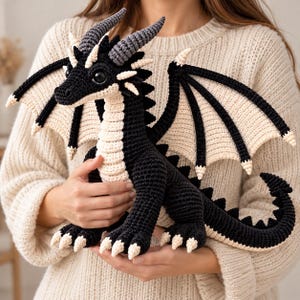 May include: A crocheted dragon plush toy featuring a black body with cream-colored underbelly, wing membranes, and claws. The creature has large dark eyes, textured horns, and a long tail with a spiked ridge along its back and neck.