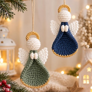 May include: Two crocheted angel ornaments hanging by twine. One angel features a sage green dress, the other a navy blue dress. Both have white heads, white wings, and gold halos with matching gold trim along the bottom edges of their dresses.
