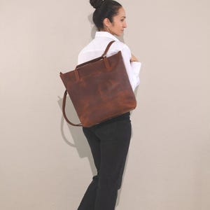 Peut inclure: Un grand sac fourre-tout rectangulaire en cuir marron foncé avec deux anses supérieures et une longue bandoulière. Le sac présente une finition mate et lisse avec des variations de tons visibles dans le cuir, une base plate et un design ouvert.