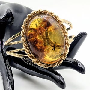 Op de afbeelding: Een goudkleurige armband met een grote, ovaalvormige barnsteen als middelpunt. De barnsteen is doorschijnend met zichtbare donkere organische insluitingen en is omlijst door een decoratieve, gedraaide touwachtige metalen rand.