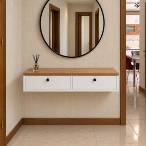 Peut inclure: Une console murale flottante avec un plateau en bois clair et deux tiroirs blancs dotés de petits boutons noirs. Au-dessus de la console se trouve un grand miroir circulaire avec un cadre noir fin. Un diffuseur à roseaux en verre est posé.