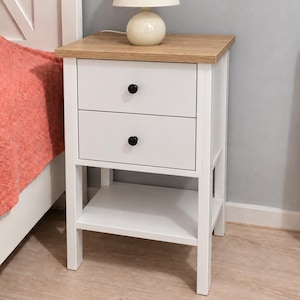 May include: A white bedside table with a light wood-toned top, featuring two drawers with small black round knobs and an open bottom shelf. A small white lamp with a rounded base and a conical shade sits on the top surface of the table.