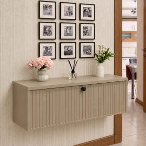 Op de afbeelding: Een aan de muur gemonteerde beige kast met een verticaal geribbeld voorpaneel en een enkele zwarte ronde knop. Bovenop staan een witte vaas met roze bloemen, een glazen geurstokjeshouder met zwarte stokjes en een witte vaas met witte bloemen.