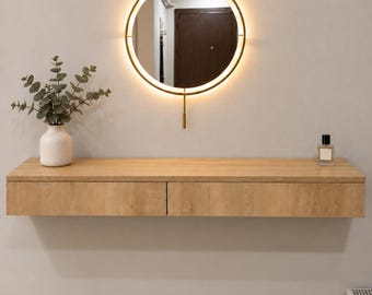 May include: A light wood floating console table with two drawers mounted on a wall. Above the table is a circular mirror with a gold frame and integrated LED backlighting. A white vase with green eucalyptus stems and a small glass bottle sit on top.
