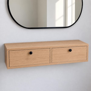 May include: A light oak wood floating wall shelf featuring two rectangular drawers. Each drawer is equipped with a single small, round, matte black knob. The shelf has a clean, horizontal design with a visible wood grain texture throughout the surface.