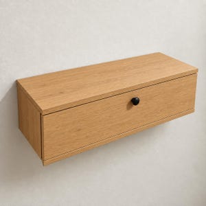 May include: A rectangular wall-mounted floating shelf featuring a light oak wood grain finish. The unit includes a single front-facing drawer with a small, round, matte black knob centered on the drawer face, providing a clean and functional storage solution.