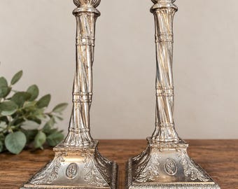 Puede incluir: Un par de candelabros de metal plateado con un diseño de columna retorcida, una parte superior acampanada y una base cuadrada decorada con grabados de hojas y un pequeño emblema ovalado central en cada lado.