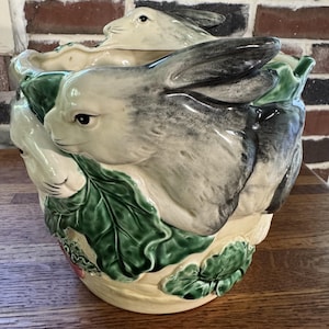 May include: A ceramic planter featuring a relief design of grey and white rabbits nestled among large, textured green leaves. The base of the planter is a cream color, and the rabbits are depicted in a resting position around the exterior.