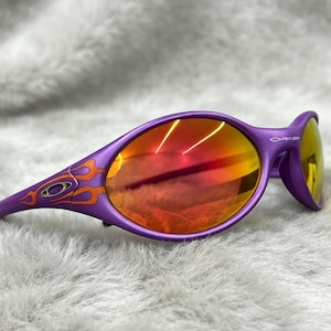 May include: Purple sunglasses featuring an oval frame with orange flame graphics on the temples. The lenses have a reflective red and orange tint. The Oakley logo is visible on the side of the frame near the hinge, showcasing a wrap-around design.