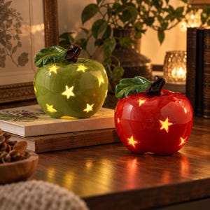 May include: Two ceramic apple-shaped lanterns, one green and one red, each featuring cut-out star shapes that emit a warm internal glow. Both apples have a glossy finish, a brown stem, and a green leaf attached to the top of the fruit structure.