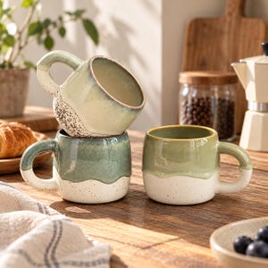 Puede incluir: Tres tazas de cerámica con un diseño bicolor que presenta una sección superior lisa y brillante en verde salvia o azul verdoso y una base mate de color blanco roto con motas. Las tazas tienen asas redondeadas y una curva definida.