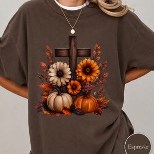 May include: A dark brown t-shirt featuring a graphic print of a wooden cross surrounded by autumn-themed flowers, orange and beige pumpkins, and fall foliage. The design includes a variety of warm-toned leaves in shades of orange, red, and brown.