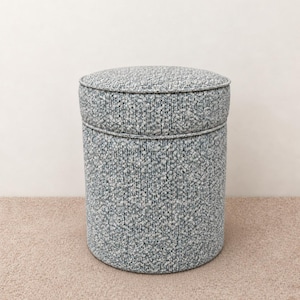 May include: A cylindrical ottoman upholstered in a textured, woven fabric featuring a speckled pattern of light blue, grey, and white threads. The piece has a distinct circular top cushion with piped edges and a straight, vertical body design.