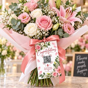 May include: A bouquet of pink and white roses, lilies, and baby's breath wrapped in pink paper with a pink ribbon. A rectangular tag hangs from the ribbon, featuring the text "Congratulations" and "Scan to hear my message!" with a QR code.