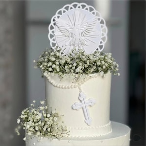 Può includere: Una torta bianca decorata con piccoli fiori bianchi, una collana di perle bianche e un ornamento a croce bianca. Un topper circolare bianco con un disegno di colomba si trova sopra la torta, circondato da un bordo con motivo a pizzo.
