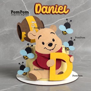 May include: A cake topper set featuring a light brown bear character in a red shirt holding a large yellow letter D. The set includes a brown honey pot with a yellow label, several small black and yellow bees with light blue wings, and the name Daniel.