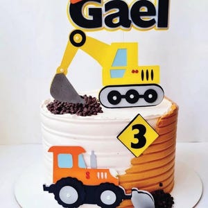 Puede incluir: Un pastel redondo decorado con glaseado blanco y naranja, con un adorno de excavadora amarilla con el nombre Gael en letras negras, un letrero amarillo en forma de diamante con el número 3 y un recorte de tractor naranja con chispas de chocolate.