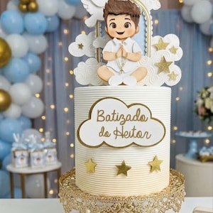 Puede incluir: Un pastel cilíndrico blanco con estrías horizontales, coronado con una figura de niño en ropa blanca sosteniendo un rosario. Un letrero en forma de nube muestra el texto Batizado do Heitor en letras doradas, decorado con estrellas doradas.