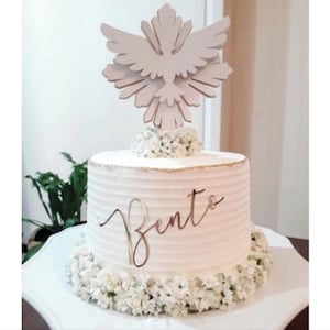 Può includere: Una torta cilindrica bianca con texture a righe orizzontali, decorata con piccoli fiori bianchi alla base e sulla sommità. Un'insegna dorata in corsivo con la scritta Bento è fissata sul lato, sormontata da un topper a forma di colomba.