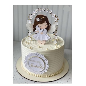 Può includere: Una torta glassata bianca con striature orizzontali, decorata con un topper circolare raffigurante un angelo dai capelli castani che tiene una colomba, incorniciato da un arco floreale bianco. Un'etichetta smerlata riporta Batizado da Isabella.