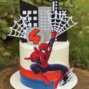 Puede incluir: Un pastel cilíndrico con glaseado blanco, con una banda roja y azul en la base. Un recorte grande de Spider-Man está centrado en el frente. Un adorno para pastel muestra un horizonte urbano negro y gris, telarañas blancas y un letrero circular con el número 4 en rojo.