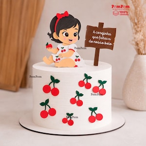 Puede incluir: Un pastel cilíndrico blanco decorado con recortes de cerezas rojas y un adorno de papel que muestra a un bebé con un lazo rojo, sosteniendo una cereza, junto a un letrero marrón con el texto A cerejinha que faltava do nosso bolo en blanco.
