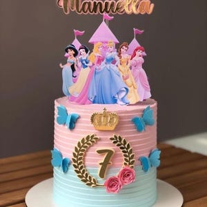 Puede incluir: Un pastel cilíndrico con glaseado rosa en la parte superior y azul claro en la inferior, con una corona dorada, un número 7 dorado dentro de una corona de laurel, cuatro mariposas azules, dos rosas rosadas y un adorno con el nombre Manuella.